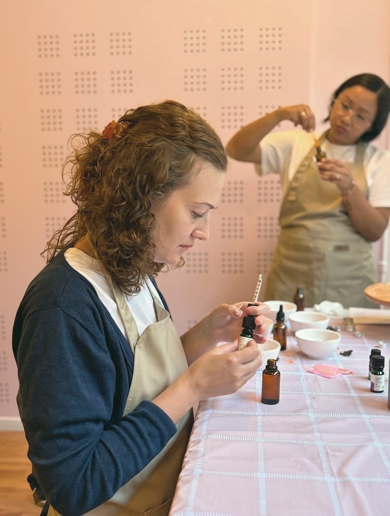 A woman measuring drops of oil into a bowl while the host guides her, against a pink wall